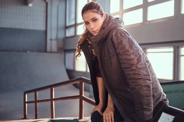 Portrait of a charming young girl with a coat covering his shoulders looking at camera while sitting on a grind rail in skatepark indoors. — Stock Photo, Image