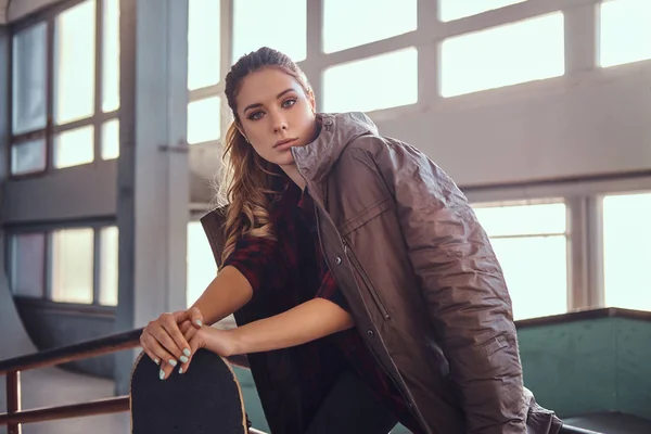 Ritratto di una bella ragazza con un cappotto che si copre le spalle appoggiato su uno skateboard e guarda la macchina fotografica mentre si siede su una grind rail in skatepark all'interno . — Foto Stock