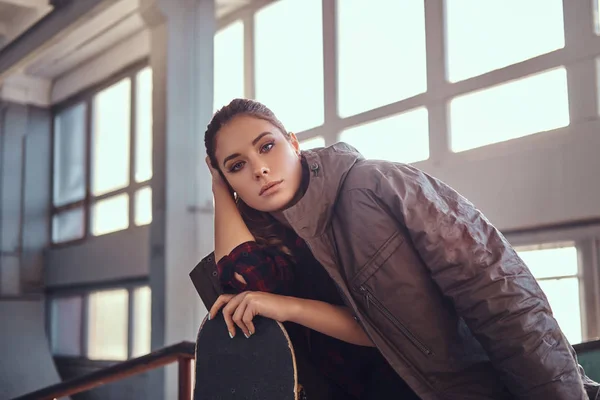 Retrato de una hermosa joven con un abrigo cubriendo sus hombros apoyado en un monopatín y mirando a la cámara mientras está sentado en un carril de molienda en el parque de skate en el interior . — Foto de Stock