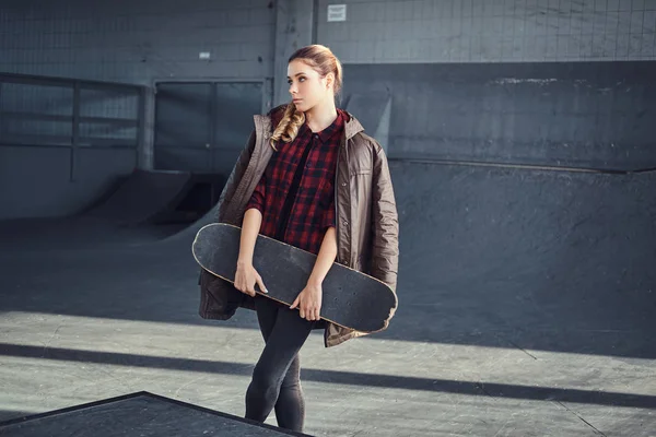 Een mooi jong meisje met een vacht die betrekking hebben op zijn schouders te houden op een skateboard in skatepark binnenshuis. — Stockfoto