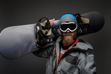 Karanlık bir arka plan üzerinde izole onun omzuna bir snowboard tutan bir tam ekipmanlı giyen kırmızı sakallı acımasız adam.