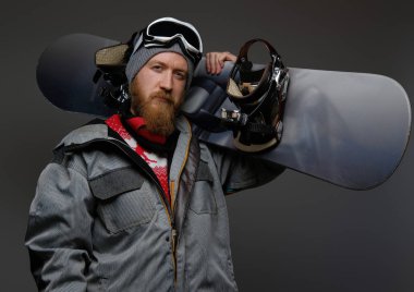 Karanlık bir arka plan üzerinde izole onun omzuna bir snowboard tutan bir tam ekipmanlı giyen kızıl sakal ile kendine güvenen adam.