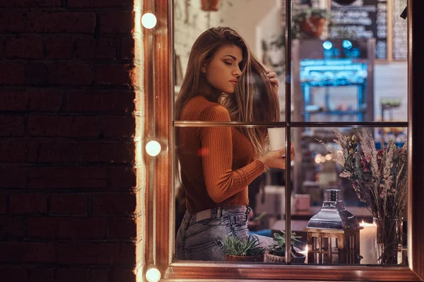Una hermosa chica con suéter sosteniendo un café mientras se sienta en un alféizar de la ventana dentro de la cafetería —  Fotos de Stock