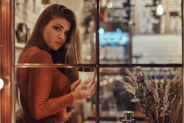 A beautiful girl wearing sweater holding a coffee while sitting on a window sill inside the cafe — Stock Photo, Image