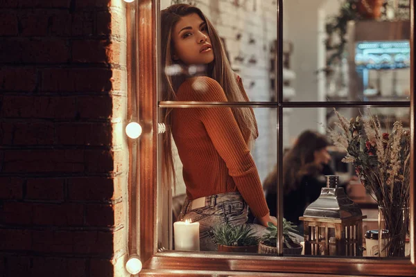 Una hermosa chica solitaria mirando a la cámara mientras está sentado en un alféizar de la ventana dentro de la cafetería — Foto de Stock