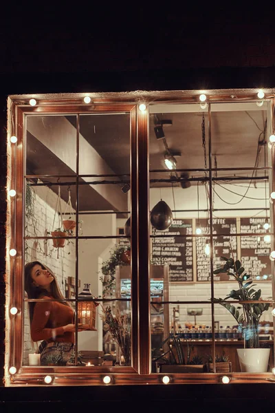 Krásná dívka, která nosí svetr drží Svíčka Lucerna zatímco sedí na okenním parapetu uvnitř kavárny — Stock fotografie