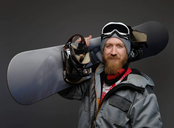 Selbstbewusster Mann mit rotem Bart und voller Ausrüstung mit einem Snowboard auf der Schulter, lächelnd und in eine Kamera blickend, isoliert auf dunklem Hintergrund. — Stockfoto