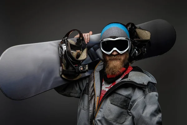 Brutální muž s červeným vousem nosí plné vybavení drží na snowboardu na rameni, izolované na tmavém pozadí. — Stock fotografie