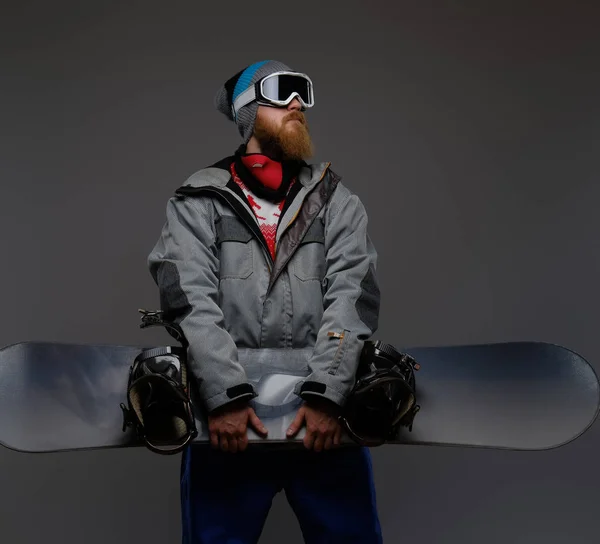 Homem brutal com uma barba vermelha vestindo um equipamento completo segurando um snowboard e olhando para longe, isolado em um fundo escuro . — Fotografia de Stock