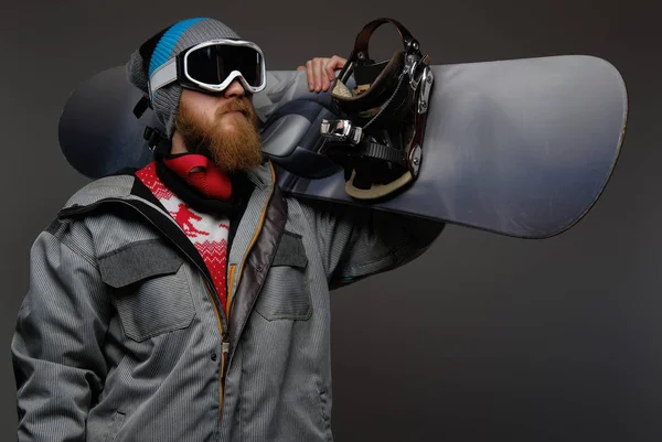 Brutalny mężczyzna z czerwoną brodą sobie pełne wyposażenie gospodarstwa na snowboardzie na ramieniu, na białym tle na ciemnym tle. — Zdjęcie stockowe