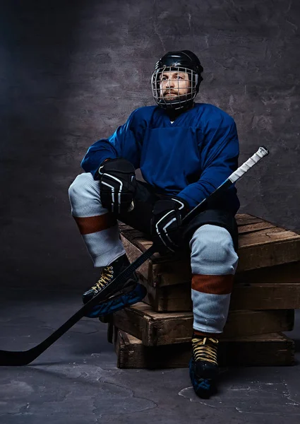 Retrato de um jogador de hóquei barbudo vestindo equipamento desportivo completo segurando uma vara de hóquei enquanto sentado em paletes de madeira . — Fotografia de Stock