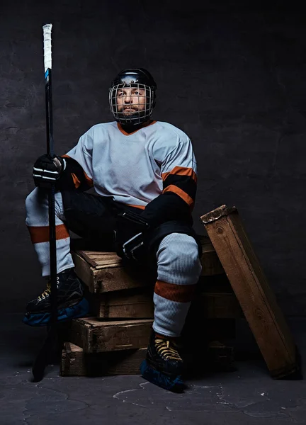 Retrato de um jogador de hóquei barbudo vestindo equipamento desportivo completo segurando uma vara de hóquei enquanto sentado em paletes de madeira . — Fotografia de Stock