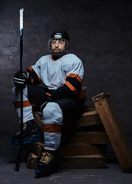 Retrato de um jogador de hóquei barbudo vestindo equipamento desportivo completo segurando uma vara de hóquei enquanto sentado em paletes de madeira . — Fotografia de Stock