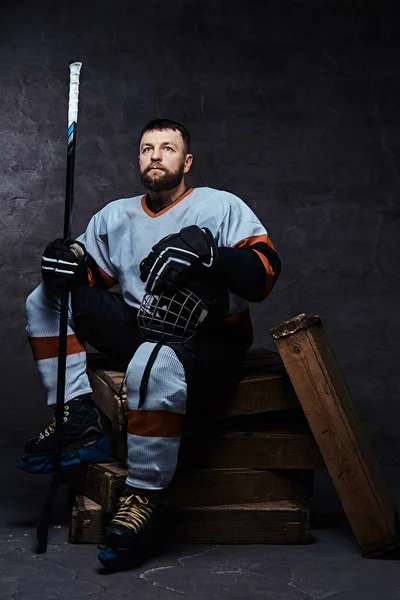 Retrato de um jogador de hóquei barbudo vestindo equipamento desportivo completo segurando um bastão de hóquei e capacete enquanto sentado em paletes de madeira . — Fotografia de Stock