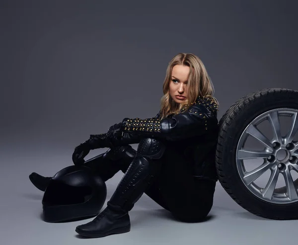 Mode, sport, extrême. Sensuelle motard fille portant des vêtements de moto assis sur un sol avec roue de voiture dans un studio . — Photo