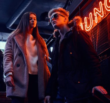 Trendy yeraltı gece kulübü için merdiven, arkadan aydınlatmalı tabela üzerinde duran genç bir çift arka planda giyinmiş.