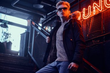 Kürk başlık ve yeraltı gece kulübü için merdiven, arka plan arkadan aydınlatmalı tabela Instalar güneş gözlüğü takan paltoyla kendine güvenen genç bir adam