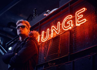 Kürk başlık ile bir kat ve kollarını ile güneş gözlüğü giyen genç bir kendine güvenen adam ayakta geçti yeraltı gece kulübü için merdiven, arka plan arkadan aydınlatmalı tabela