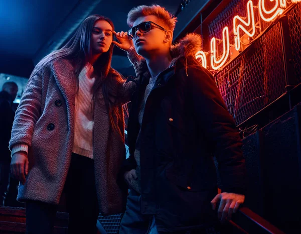 Una pareja joven y elegante con ropa de abrigo de pie en la escalera a la discoteca subterránea, un letrero retroiluminado en el fondo —  Fotos de Stock