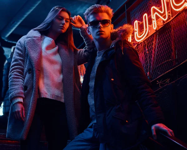 Una pareja joven y elegante con ropa de abrigo de pie en la escalera a la discoteca subterránea, un letrero retroiluminado en el fondo —  Fotos de Stock