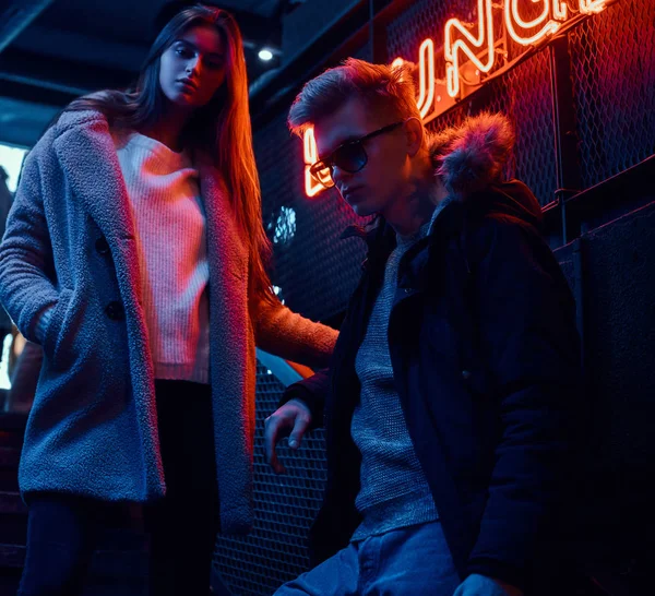 Młoda para stylowe nosić ciepłe ubrania, stojąc na schody do podziemnego klubu nocnego, szyld podświetlany w tle — Zdjęcie stockowe