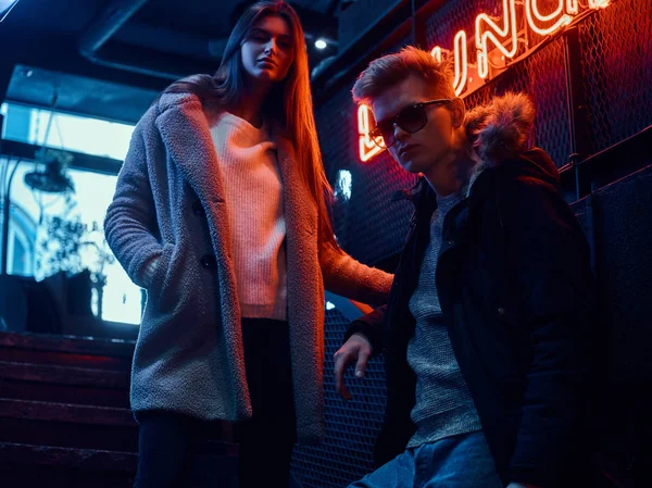 Trendy gekleed jonge paar staande op de trap naar de ondergrondse nachtclub, een verlicht bord op de achtergrond — Stockfoto