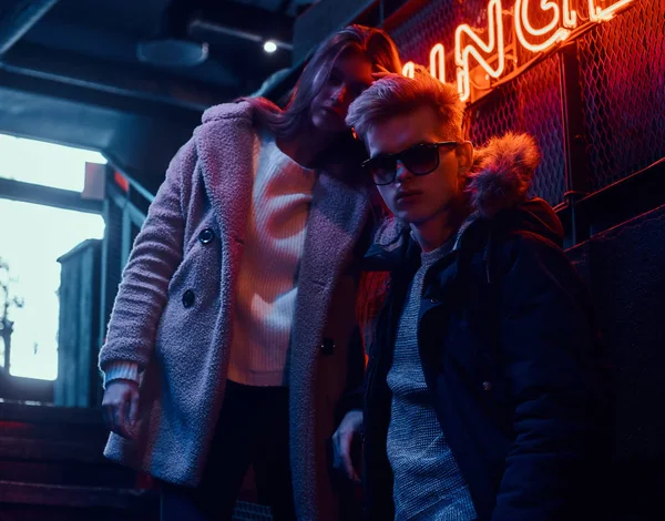 Trendy yeraltı gece kulübü için merdiven, arkadan aydınlatmalı tabela üzerinde duran genç bir çift arka planda giyinmiş.
