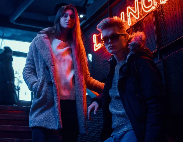 Um jovem casal elegante vestindo roupas quentes em pé na escada para a discoteca subterrânea, uma placa retroiluminada no fundo — Fotografia de Stock