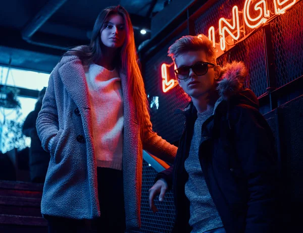 Moda vestido jovem casal de pé na escada para a discoteca subterrânea, uma placa retroiluminada no fundo — Fotografia de Stock
