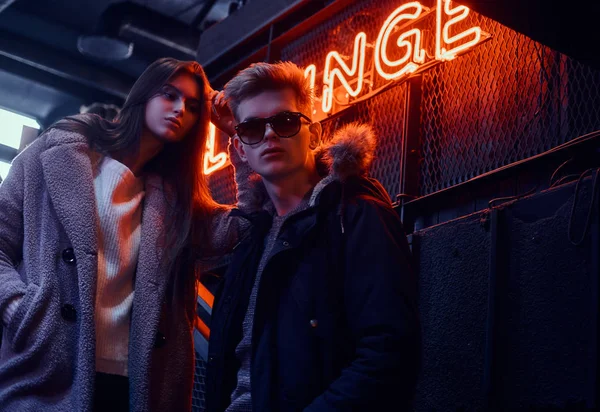 Ein junges stylisches Paar in warmer Kleidung steht auf der Treppe zum Nachtclub, im Hintergrund ein hinterleuchtetes Schild — Stockfoto