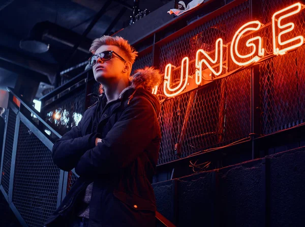 Een jonge vertrouwen kerel het dragen van een jas met bont capuchon en zonnebril met zijn armen gekruist staande op de trap naar de ondergrondse nachtclub, een verlicht bord op de achtergrond — Stockfoto