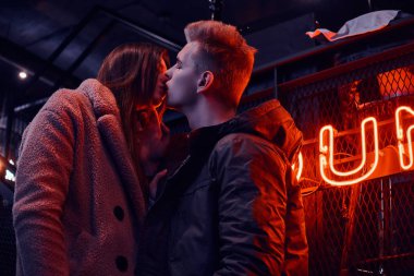 Trendy tutkulu genç çift öpüşme sırasında ayakta endüstriyel iç kafede, arka plan arkadan aydınlatmalı tabela giyinmiş