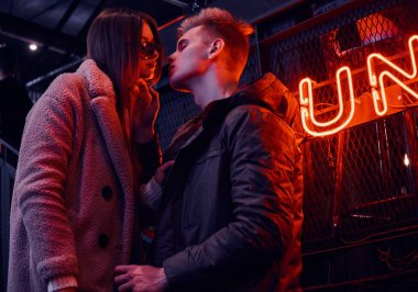 Trendy tutkulu genç çift öpüşme sırasında ayakta endüstriyel iç kafede, arka plan arkadan aydınlatmalı tabela giyinmiş