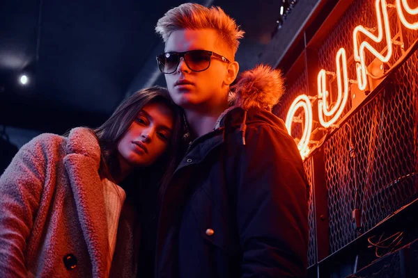 Um jovem casal elegante vestindo roupas quentes em pé no café com interior industrial, uma placa retroiluminada no fundo — Fotografia de Stock