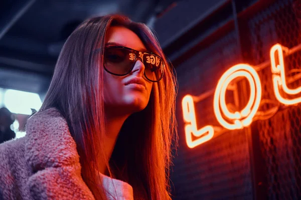 Retrato de cerca de una sensual joven con gafas de sol y abrigo de pie en la cafetería con el interior industrial, un letrero retroiluminado en el fondo —  Fotos de Stock