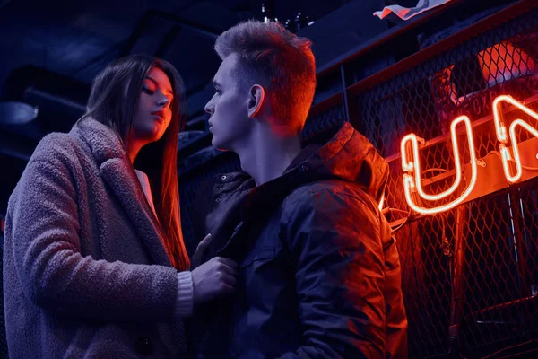 Genç bir şık çift tutkuyla bak birbirlerine ayakta endüstriyel iç kafede, arka plan arkadan aydınlatmalı tabela