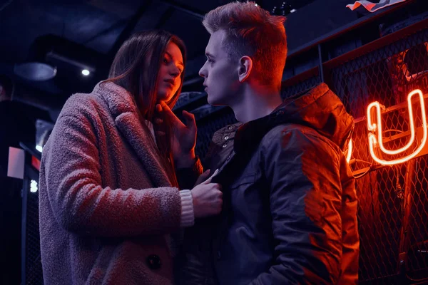 Genç bir şık çift tutkuyla bak birbirlerine ayakta endüstriyel iç kafede, arka plan arkadan aydınlatmalı tabela