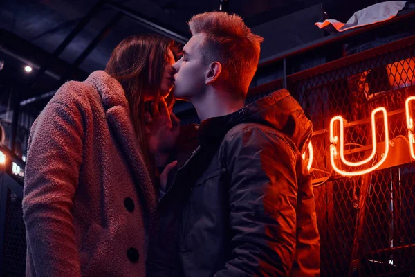 Stilvolles junges Paar blickt sich leidenschaftlich an, während es im Café mit industriellem Interieur steht, im Hintergrund ein hinterleuchtetes Schild — Stockfoto