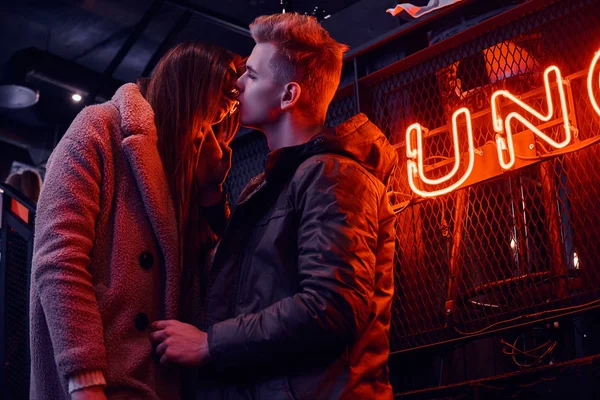 Moda vestida pareja joven apasionada besándose mientras está de pie en la cafetería con interior industrial, un letrero retroiluminado en el fondo —  Fotos de Stock