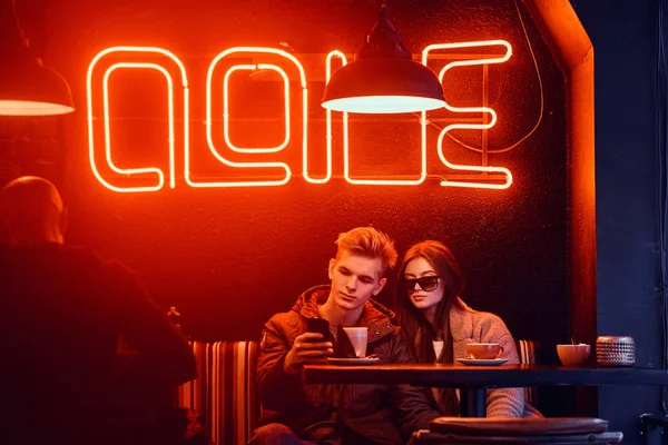 Trendy oblečený mladý pár sedí v kavárně s průmyslový interiér, chatování s přáteli nebo sledovat něco, co na telefonu — Stock fotografie