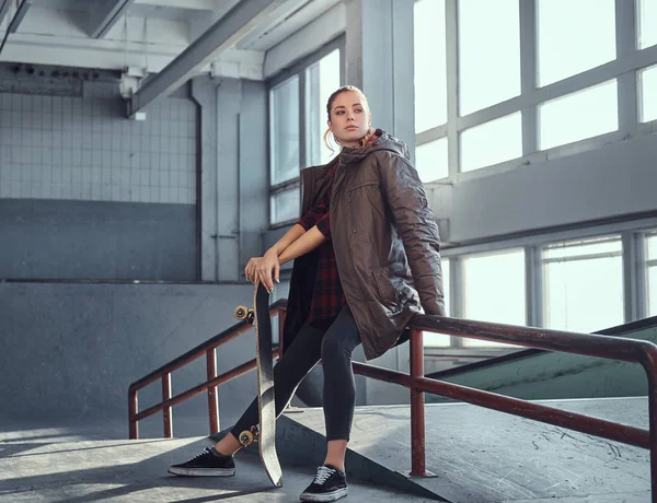 Uma linda menina vestindo uma camisa quadriculada e casaco com seu skate sentado em um trilho de moagem no parque de skate dentro de casa . — Fotografia de Stock