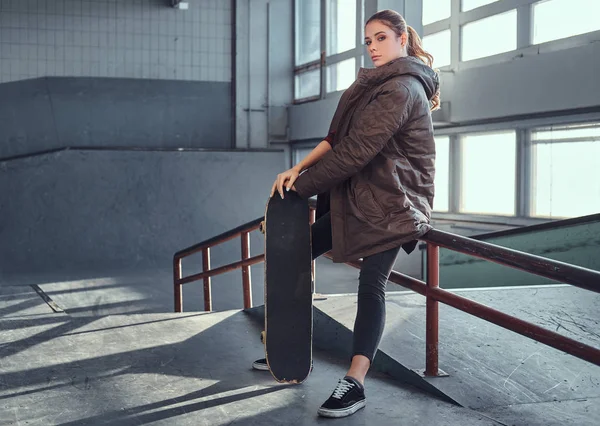 Una hermosa joven con una camisa a cuadros y un abrigo con su monopatín sentado en un carril de molienda en el parque de skate en el interior . — Foto de Stock