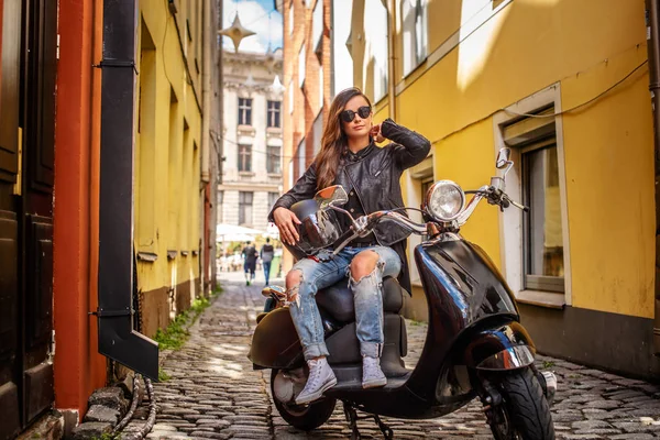 Modisches Mädchen mit Sonnenbrille, Lederjacke und zerrissenen Jeans sitzt auf einem schwarzen Oldtimer-Roller in einer alten engen Straße. — Stockfoto