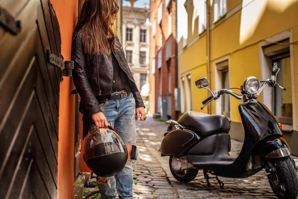 时尚女孩拿着防护头盔, 靠在一辆黑色摩托车附近的墙上, 站在一条狭窄的老街道上. — 图库照片