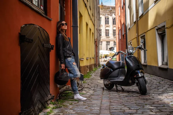 Modieuze meisje houden een beschermende helm terwijl leunend op een muur in de buurt van een zwarte scooter, staande op een oude smalle straat. — Stockfoto