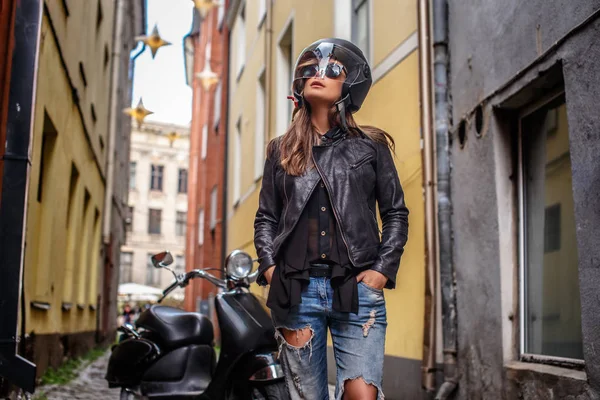 Ragazza alla moda in casco protettivo e occhiali da sole indossando una giacca di pelle e jeans strappati in piedi vicino a uno scooter nero su una vecchia strada stretta . — Foto Stock