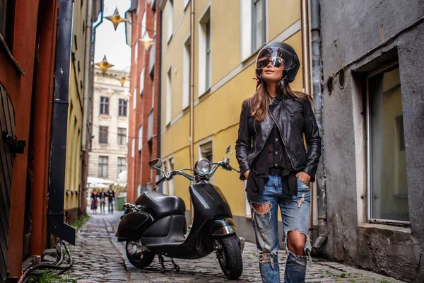 Menina na moda em capacete protetor e óculos de sol usando uma jaqueta de couro e jeans rasgado em pé perto de uma scooter preta em uma rua estreita velha . — Fotografia de Stock