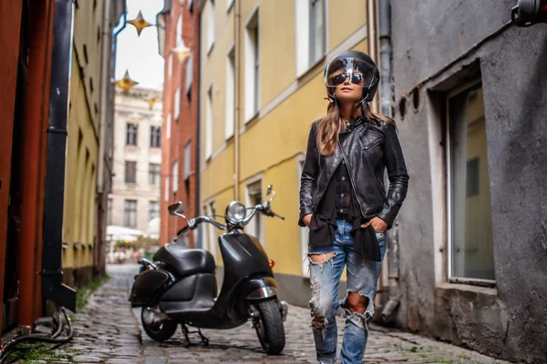 Modieuze meisje in beschermende helm en zonnebril dragen van een leren jas en gescheurde jeans permanent in de buurt van een zwarte scooter op een oude smalle straat. — Stockfoto