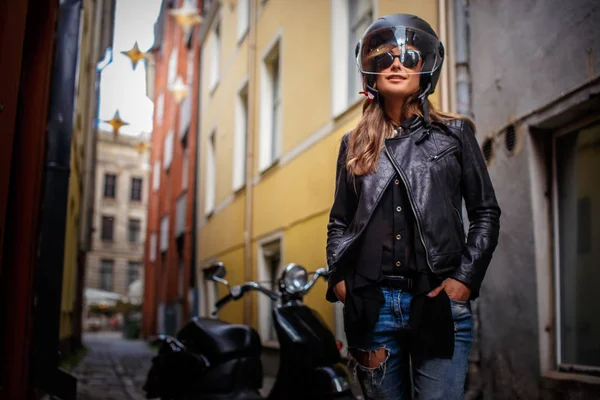 Modisches Mädchen mit Schutzhelm und Sonnenbrille in Lederjacke und zerrissenen Jeans, das in der Nähe eines schwarzen Motorrollers in einer alten engen Straße steht. — Stockfoto