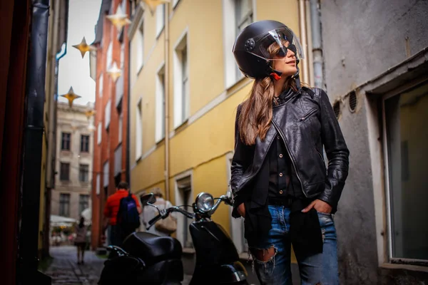 Modieuze meisje in beschermende helm en zonnebril dragen van een leren jas en gescheurde jeans permanent in de buurt van een zwarte scooter op een oude smalle straat. — Stockfoto
